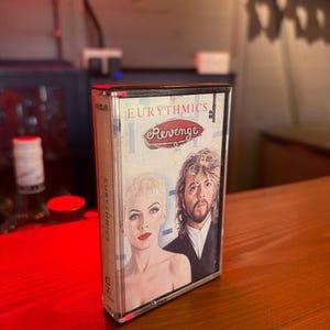 Può includere: Una musicassetta vintage dell'album "Revenge" degli Eurythmics. La copertina dell'album presenta un ritratto dei membri della band. La cassetta è in una custodia di plastica trasparente, con il logo RCA visibile.