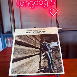 May include: A vintage vinyl record album cover for Don Williams' "You're My Best Friend" displayed on a decorative black metal stand. The album cover features a photograph of a man in a denim jacket and jeans. A neon sign that says "Longdog's" is in the background.