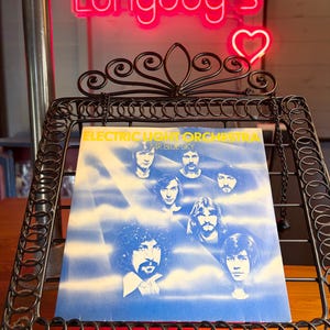 May include: A vinyl record cover for Electric Light Orchestra's Mr. Blue Sky, featuring portraits of the band members against a blue and white cloudy background, displayed on a black metal wire stand with decorative scrollwork at the top.
