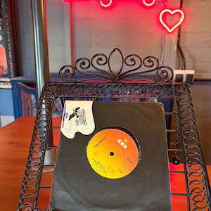 May include: A black metal record holder displaying a vinyl record by Tina Charles. The record label is yellow and orange with the text "Tina Charles" and "I Love to Love". The record is in a black sleeve, and the holder is in front of a neon sign that says "Longdog's".