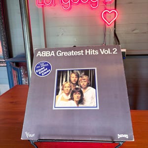 Könnte beinhalten: Albumcover von ABBA Greatest Hits Vol. 2 mit einem Bandfoto. Das Album steht auf einem schwarzen Metallständer. Das pinkfarbene Neonzeichen darüber zeigt "Longdog's".