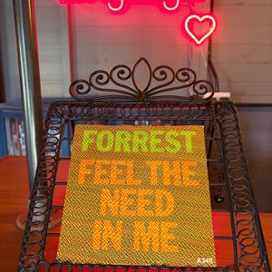 May include: A black metal stand displays a sign with the text "FORREST FEEL THE NEED IN ME" in green and orange. Above, a neon sign reads "Longdog's" with a heart symbol. The stand features a decorative, scrolled design.