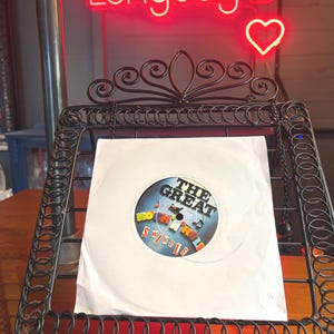 May include: A white vinyl record sleeve featuring a circular label with the text THE GREAT ROCK 'N' ROLL SWINDLE in black and colourful block letters, displayed on a black metal wire stand with decorative scrollwork at the top.