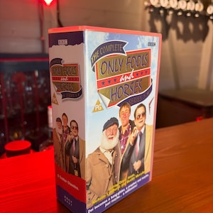 Pode incluir: Caixa de DVD da série de comédia britânica "Only Fools and Horses". A caixa apresenta o título do programa em letras douradas sobre um fundo azul, com imagens dos personagens principais. O logotipo da BBC é visível.