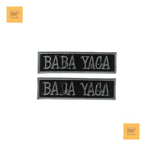 Può includere: Due toppe rettangolari nere con la scritta "BABA YAGA" ricamata in grigio chiaro. Le toppe sono affiancate, con il testo centrato su ogni toppa. Le toppe sono probabilmente per abbigliamento o accessori.