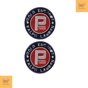 Può includere: Due toppe circolari con un centro rosso e una "P" bianca con un piccolo disegno della bandiera americana. Le toppe sono circondate da un bordo blu navy con le parole "RALPH LAUREN" e "EST. 1967" in bianco.