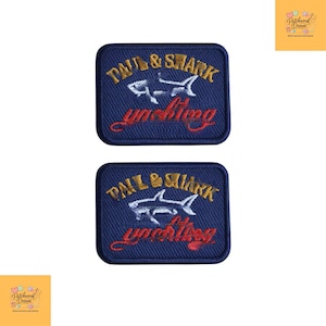 Könnte beinhalten: Zwei rechteckige, dunkelblaue Stickaufnäher mit dem Text Paul & Shark in Goldfaden über einer weißen Haifisch-Silhouette und dem Wort yachting in roter Schreibschrift. Die Aufnäher haben abgerundete Ecken und einen dunkelblauen Rand.