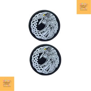 May include: Two circular embroidered patches featuring a white raven design with intricate knotwork patterns on a black background. Each raven has a single yellow eye and detailed feather textures, enclosed within a solid black circular border.
