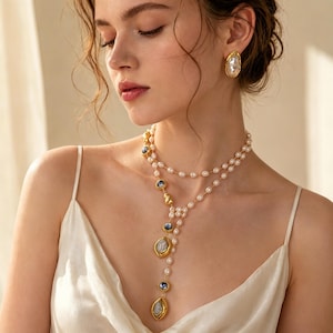 Puede incluir: Un collar dorado y pendientes a juego con perlas y piedras preciosas azules. El collar tiene un diseño en capas con un colgante central. Los pendientes tienen una perla en el centro. La joyería se usa con un vestido blanco.