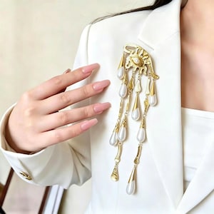 May include: A gold-coloured brooch with a decorative design, featuring dangling teardrop-shaped elements and faux pearls. The brooch is pinned to a white blazer, showcasing its intricate details and elegant style.
