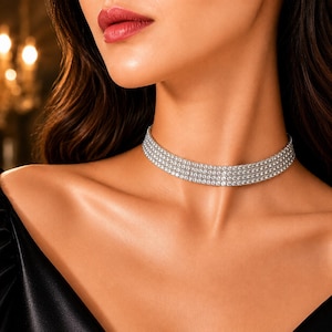 Può includere: Una scintillante collana girocollo argentata con più file di cristalli trasparenti a taglio quadrato. La collana è indossata attorno al collo, riflettendo la luce. La modella indossa un top nero.