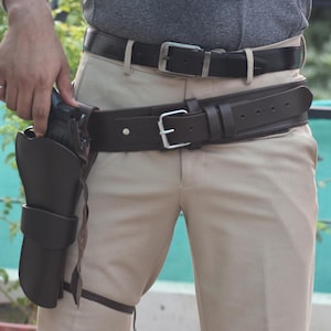 May include: A dark brown leather holster with a gun, attached to a brown leather belt. The belt is worn over beige trousers. A second black leather belt is visible above the brown belt. The person is wearing a grey shirt.