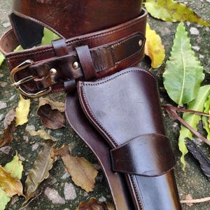 Op de afbeelding: Een bruine leren pistoolholster- en riemset. De riem heeft een messing gesp en lussen voor kogels. De holster is ontworpen om een ​​handvuurwapen te bevatten. De set is gemaakt van donkerbruin leer en lijkt handgemaakt.
