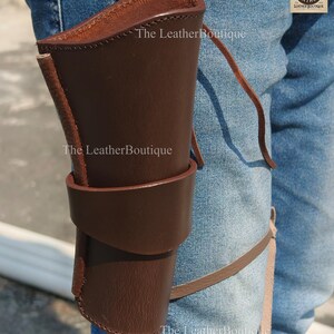 Op de afbeelding: Een bruin leren holster met een riemlus en beenband. De holster is ontworpen om een vuurwapen te bevatten en is gemaakt van stevig leer. Het logo The Leather Boutique is zichtbaar.