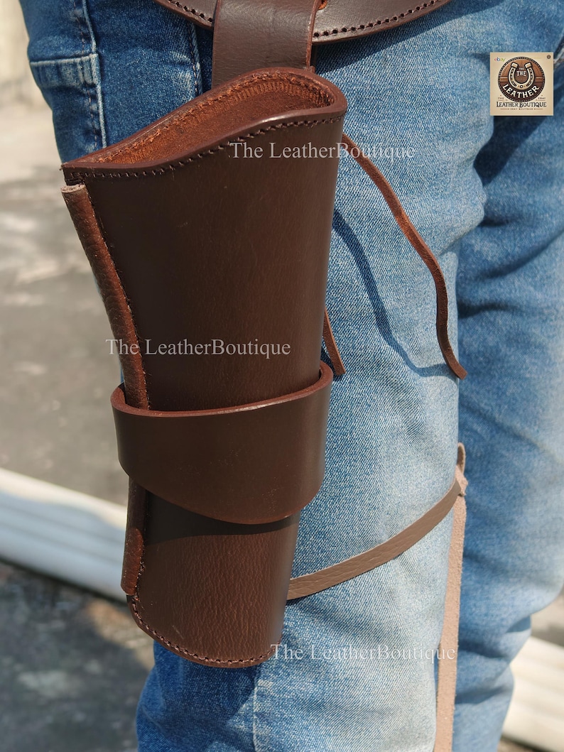 Op de afbeelding: Een bruin leren holster met een riemlus en beenband. De holster is ontworpen om een vuurwapen te bevatten en is gemaakt van stevig leer. Het logo The Leather Boutique is zichtbaar.