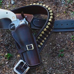 Peut inclure: Un revolver argenté avec une poignée couleur crème repose dans un étui en cuir marron foncé. L'étui est fixé à une ceinture en cuir assortie comportant une série de cartouches à douille en laiton dans des boucles et une boucle en métal.