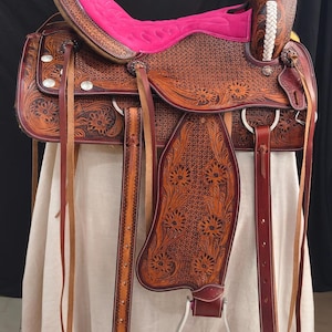 May include: A brown leather western saddle with intricate floral tooling and a bright pink suede seat. The saddle features silver conchos, stirrups, and a matching headstall. The design includes a decorative horn and a white braided accent.