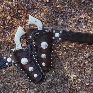 May include: A dark brown leather gun belt featuring two holsters, each containing a revolver with a white grip. The belt and holsters are decorated with silver-toned metal conchos and studs, and the belt includes a silver-toned metal buckle.