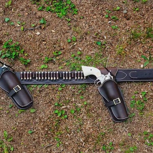 Peut inclure: Une ceinture de pistolet de style western avec deux étuis en cuir marron, chacun contenant un revolver à manche blanc. La ceinture est en cuir noir avec des coutures décoratives et une boucle argentée. Une rangée de balles est présente.