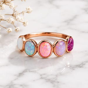 Op de afbeelding: Een roségouden ring met vijf ovale cabochonstenen op een rij. De stenen vertonen een kleurverloop, van wit aan de linkerkant naar lichtblauw, felroze, zacht lavendel en diep magenta aan de rechterkant.