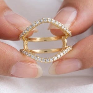 Peut inclure: Une bague en or au design à double anneau avec une silhouette incurvée et ouverte. Les bandes supérieure et inférieure sont incrustées de petites pierres claires taillées en rond qui suivent le contour ondulé du métal, créant un aspect superposé.