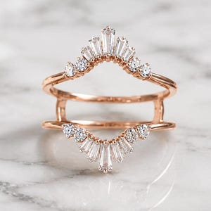 Puede incluir: Un protector de anillo en oro rosa con dos bandas curvas conectadas por barras verticales. Los arcos superior e inferior están engastados con una combinación de piedras transparentes en talla baguette y redonda en abanico.