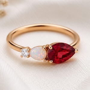 Op de afbeelding: Een gouden ring met een peervormige rode edelsteen, een ronde witte opaal en een kleine ronde heldere diamant. De stenen zijn op een rij gezet langs de dunne gouden band, wat zorgt voor een verfijnd arrangement van gekleurde edelstenen.