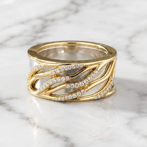 May include: A wide band ring featuring a two-tone design with a polished silver-colored inner band and a yellow gold-colored outer layer. The outer layer has an openwork wave pattern embellished with small, clear, round-cut stones.