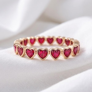 Könnte beinhalten: Ein goldener Ring mit einer durchgehenden Reihe kleiner, facettierter, herzförmiger roter Steine, die in einem sich wiederholenden Muster um den gesamten Umfang des Schmuckstücks angeordnet sind und ein lebendiges, symmetrisches Design bilden.