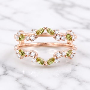 Könnte beinhalten: Ein Ring-Guard aus Roségold mit zwei parallelen Bändern, die durch seitliche Stege verbunden sind. Das Design ist mit marquise-geschliffenen grünen Peridoten und runden, weißen Brillanten in einem symmetrischen, blattartigen Muster besetzt.