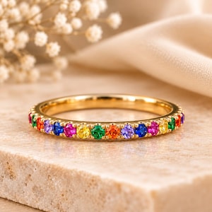 May include: A gold band ring featuring a continuous row of small, round, multi-colored gemstones in a rainbow sequence. The stones include red, orange, yellow, green, blue, and purple hues set into the polished metal band in a prong setting.