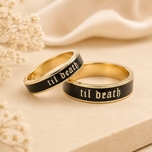 May include: Two gold-toned metal rings featuring a black enamel inlay around the outer band. Each ring is engraved with the text 'til death' in a gothic-style font. The rings have a smooth, polished finish and a flat, uniform band profile.