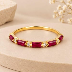 Puede incluir: Un anillo de oro con una serie de rubíes rojos rectangulares de corte baguette alternados con pequeños diamantes blancos redondos engastados en la banda. El anillo de metal tiene un acabado de oro amarillo pulido de aspecto brillante.