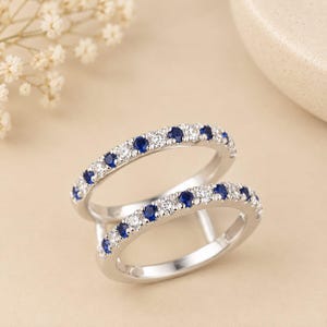 May include: A silver-toned double band ring featuring two parallel rows of alternating round-cut blue sapphires and clear white diamonds. The metal bands are polished and connected, creating a unified piece of jewelry with a sparkling finish.