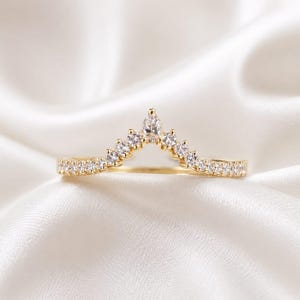 May include: A gold V-shaped ring featuring a central pear-cut clear stone flanked by a series of round-cut clear stones along the band. The metal band has a polished finish and the stones are set in a symmetrical arrangement across the top arc.