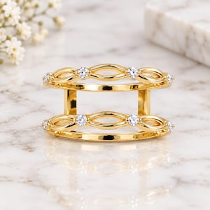 Puede incluir: Un anillo doble de oro con dos bandas paralelas conectadas por barras verticales. Cada banda presenta un patrón abierto de eslabones en forma de infinito, acentuado con pequeñas piedras redondas transparentes engastadas.