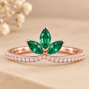 Puede incluir: Un anillo de oro rosa con una banda en forma de V engastada con pequeñas piedras blancas redondas. El centro del anillo muestra tres piedras verdes de talla marquesa dispuestas en abanico, creando una apariencia de corona.