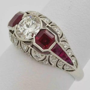 Puede incluir: Un anillo de estilo vintage con una gran piedra central de diamante transparente y redondo, flanqueado por dos rubíes cuadrados y pequeños acentos de rubí. El anillo tiene una banda de plata detallada y ornamentada con pequeños adornos de diamantes.