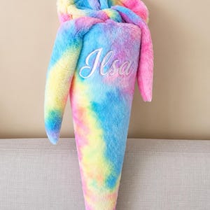 Könnte beinhalten: Eine kegelförmige Schultüte aus weichem, flauschigem Kunstfell mit einem lebendigen Batikmuster in Blau-, Rosa- und Gelbtönen. Der obere Teil verfügt über eine kapuzenartige Klappe, und der Name Ilsa ist in weißer Schreibschrift aufgestickt.