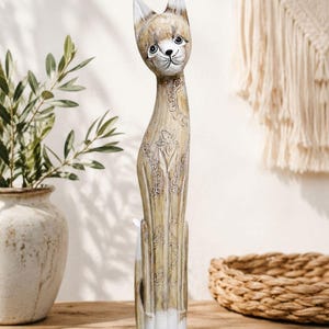 May include: A tall, wooden cat figurine with a slender body and a long neck. The cat is painted in shades of beige and white, with intricate floral patterns carved into its surface. The cat is approximately 12 inches tall and sits on a wooden surface.