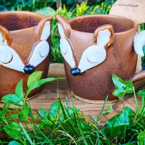Könnte beinhalten: Zwei braune Keramikbecher in Fuchsform, jeweils mit weißen Gesichtsmerkmalen, spitzen Ohren und einer kleinen schwarzen Nase. Die Becher haben geschwungene Henkel und eine glatte, glasierte Oberfläche, die das Gesicht eines Fuchses darstellt.