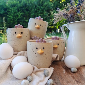 Op de afbeelding: Vier gespikkelde keramische bekers in de vorm van kippen met kleine gele snavels, donkere ogen en paarse kammen. De bekers zijn gerangschikt met verschillende witte eieren op een lichtgekleurde doek, vergezeld door een witte kan met droogbloemen.
