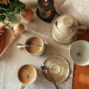 Könnte beinhalten: Draufsicht auf einen Tisch mit Kaffeetassen, einer Teekanne und einer Untertasse. Die Tassen und die Teekanne sind gesprenkelt beige. Ein Holzbrett hält eine Brezel. Eine brennende grüne Kerze und dekorative Holzobjekte sind ebenfalls vorhanden.