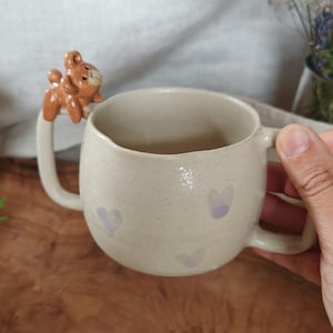 Op de afbeelding: Een beige keramische mok met een gespikkelde afwerking en lichtpaarse hartjespatronen. De mok heeft twee oren, waarvan er één is bekroond met een klein, gebeeldhouwd bruin berenfiguurtje. Het ontwerp is rond met een gladde, glanzende glazuur.