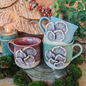 Puede incluir: Tres tazas de cerámica con grupos de setas esculpidas en el exterior. Una taza es roja con base crema, otra es azul claro con motas y otra es verde salvia con base crema. Cada taza tiene un asa y un diseño de setas con textura en relieve.