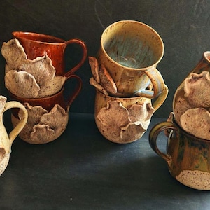 Könnte beinhalten: Eine Sammlung handgefertigter Keramikbecher mit einem einzigartigen Pilzdesign. Die Becher sind in verschiedenen Erdtönen gehalten, darunter Braun, Beige und Grün. Jeder Becher hat einen Henkel und ein dekoratives Pilzelement.