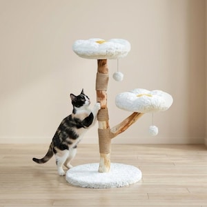 May include: A cat scratching post with two white, flower-shaped platforms and a round base. The post is made of natural wood and rope, with a small cat climbing on it. The platforms have yellow centers and dangling pom-poms.