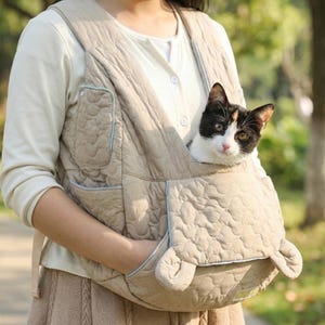 Puede incluir: Un portabebés acolchado beige con ribete azul claro, diseñado para ser usado como chaleco. Un gato blanco y negro está acurrucado dentro del portabebés. El portabebés tiene dos bolsillos y una base redondeada.