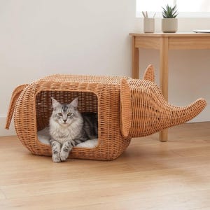 Könnte beinhalten: Ein Katzenbett aus geflochtenem Korb in Form eines Elefanten, mit einer grau-weißen Katze darin. Das Bett ist hellbraun und hat eine rechteckige Öffnung. Der Rüssel und die Ohren des Elefanten sind ebenfalls aus geflochtenem Korb.
