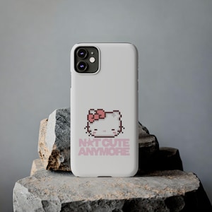 Puede incluir: Funda de teléfono blanca con un diseño de gato pixelado, lazo rosa y el texto "N*T CUTE ANYMORE" en rosa. La funda, con cámara de doble lente, se muestra sobre una roca.